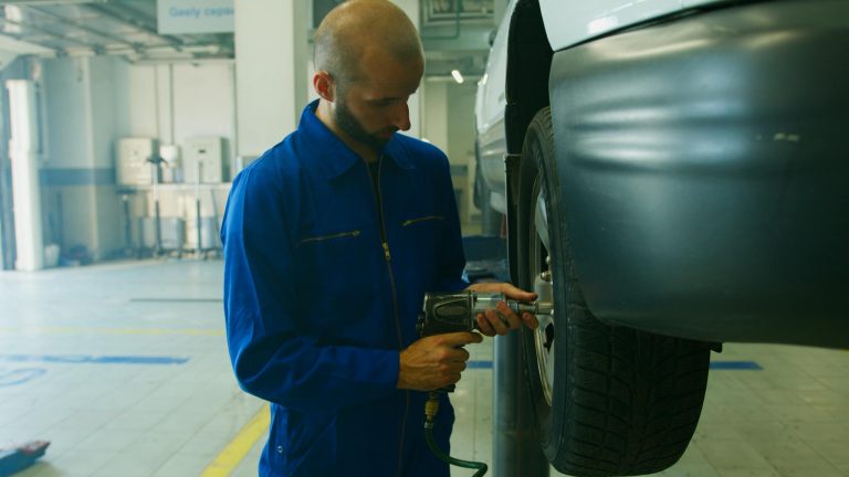 Mechanic in blue uniform servicing a car with an impact wrench in a garage.