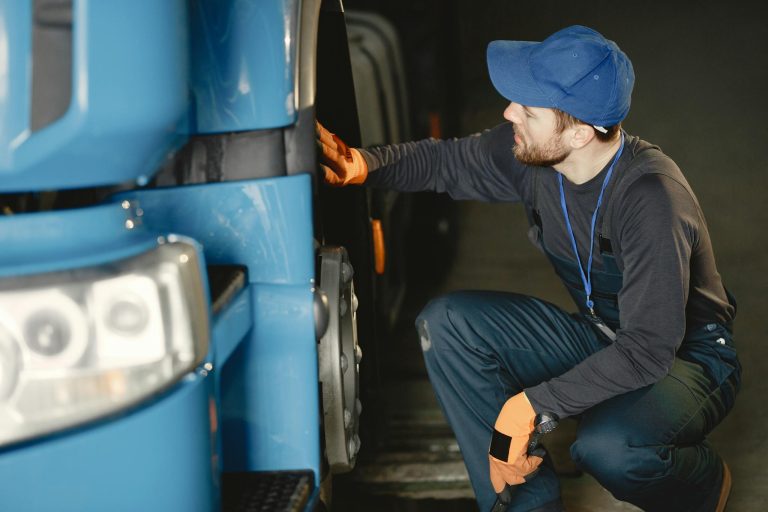 Mechanic in uniform checking a blue truck's wheel inside a garage workshop.